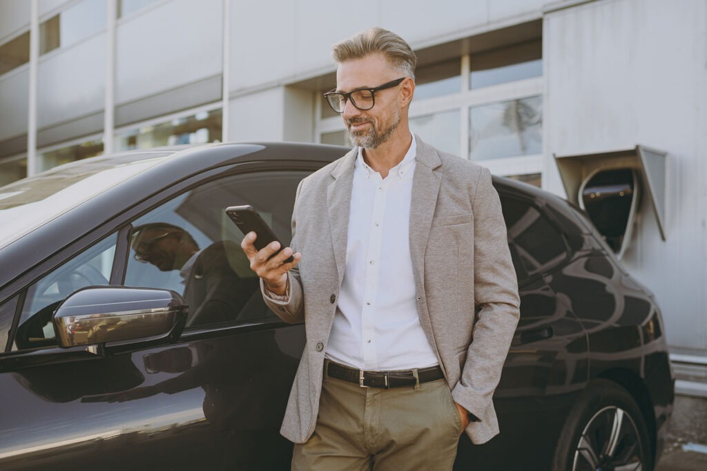 Satisfied confident adult man customer male buyer client wearing white shirt classic grey suit standing near black electric car while charging talk speak on mobile cell phone outside Business concept