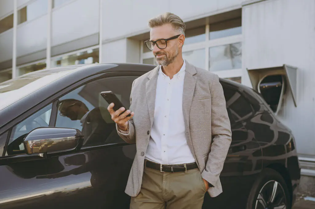 Satisfied confident adult man customer male buyer client wearing white shirt classic grey suit standing near black electric car while charging talk speak on mobile cell phone outside Business concept
